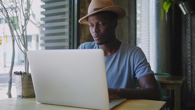 Young black university student does his homework in a busy coffee shop near his high school, works remotely on his laptop to prepare the papers due for exams