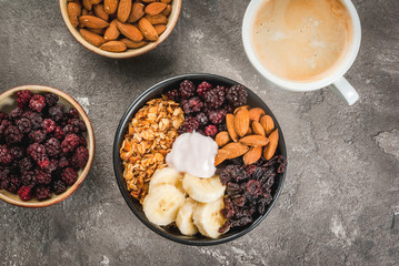Buddha bowl. Concept of healthy balanced diet breakfast: oatmeal granola, dried cherries, blackberries, banana, nuts almonds and yogurt. On a gray stone table, with cup of coffee top view copy space