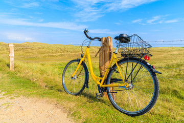 Naklejka na ściany i meble SYLT ISLAND, GERMANY - SEP 6, 2016: yellow bike parked against a field fence in countryside landscape of Sylt island near Ellenbogen beach, Germany.