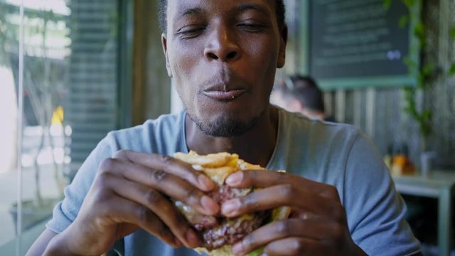 Hand held shot with selective focus of a stylish afro american man in blue shirt munching on a huge american burger with cheese and fresh vegetables.