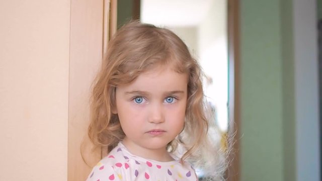 Little girl is blonde with blue eyes stands near the wall in the house. Child with regret looking at the camera,portrait of a sad girl.