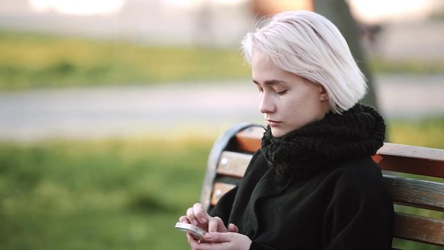 Blonde woman girl outside. Looks at the smartphone. Sits on a wooden bench. With a serious look on his face, he looks into the smartphone and plays an intellectual game. Side view.