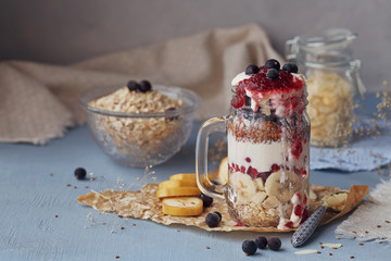Layered dessert with muesli, yogurt, banana, linen seeds, slised almonds and berries in glass mason jar on blue rustic background.