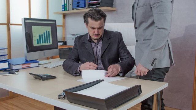A young employee is bringing a document for his boss to sign. Then the boss continues working.