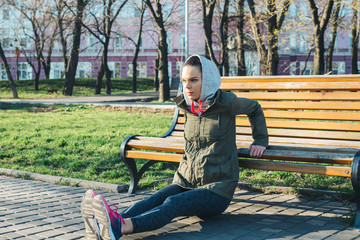 Fototapeta premium Young woman in a jacket and a hood doing exercises on triceps on a bench in the park in spring