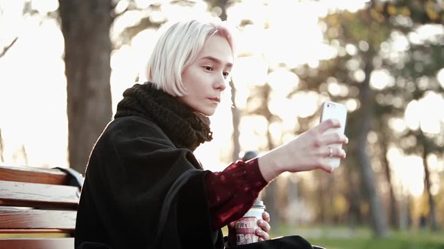 Blond woman girl outside Holds a smartphone and coffee Sits on a wooden bench Do yourself Trying to make a beautiful face The sun flashes into the camera Side view Shrugs shows tongue smiles.