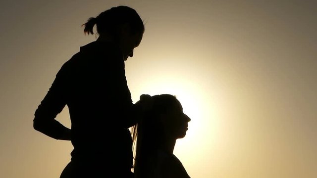 Silhouettes of Two Young Women Sitting and Putting Hair in Order at Sunset.