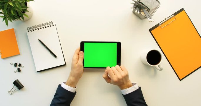 Business man using tablet device with green screen on white office desk with orange details. Male hand scrolling pages, zooming on tablet computer with green screen. Top view. Chroma key