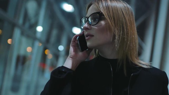 Attractive young woman in the glasses with black rim and dark clothes is near the business center in the evening. The girl is talking with someone on her smartphone. Evening in the city, good mood.