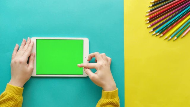 Woman using white tablet computer with green screen on blue and yellow background with colorful pencils. Female hand scrolling pages, tapping on touch screen. Chroma key. Top view