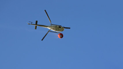 Fototapeta na wymiar Firefighting helicopter with water bucket and trail of drops seen from below against blue sky