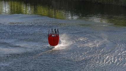 Detail of water bucket lifting out of the water under a helicopter with rotor wash on the surface of the lake