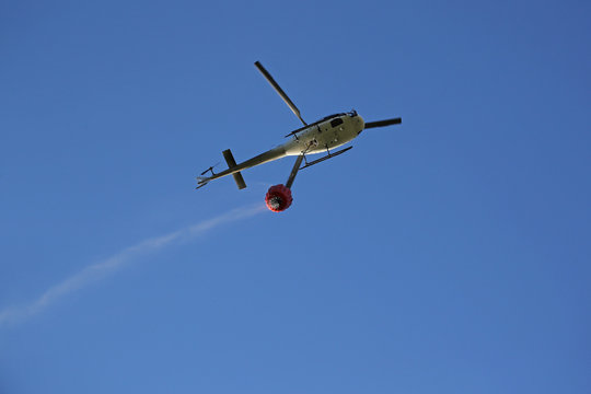 Firefighting helicopter with water bucket and trail of drops seen from below against blue sky