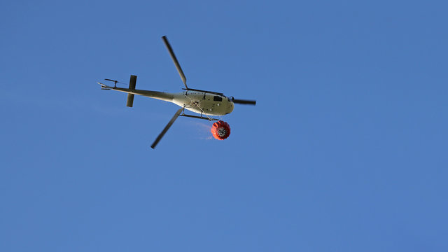 Firefighting helicopter with water bucket and trail of drops seen from below against blue sky