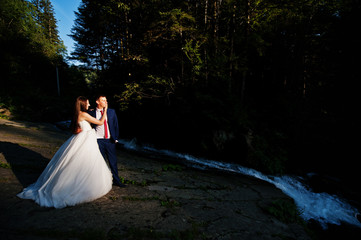 Lovely wedding couple against waterfall on sunset at Carpathian mountains.