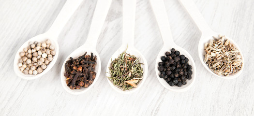 Different spices and herbs in wooden spoons on white table background. Black and white pepper, clove, savory, fennel seeds. Aromatic food cooking ingredients. Top view, closeup