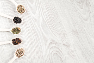 Different spices and herbs in wooden spoons on white table background. Aromatic food cooking ingredients. Black and white pepper, clove, savory, fennel seeds. Top view, free space.