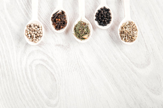 Various spices and herbs in wooden spoons on white table background. Aromatic food cooking ingredients. Black and white pepper, clove, savory, fennel seeds. Top view, free space.