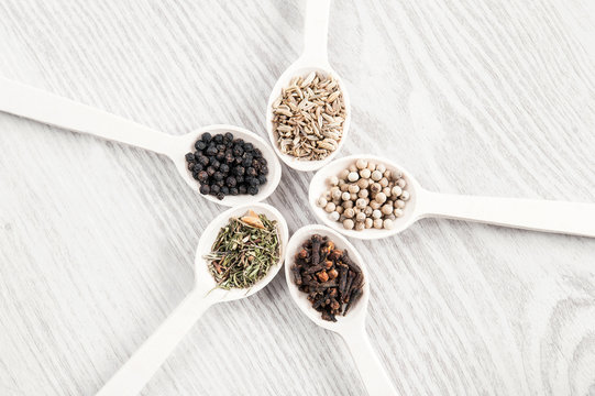 Various spices and herbs in wooden spoons on white table background. Black and white pepper, clove, savory, fennel seeds. Aromatic food cooking ingredients. Top view.