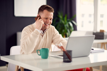 Young man working in modern office, sitting at the table with laptop, talking over mobile phone