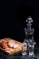 Bottle and shot glasses with vodka, slices of smoked meat and smoked sausage on wooden cutting board.