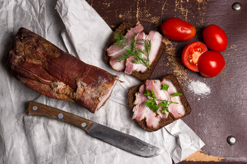 Smoked bacon with rye black bread and cuted tomatoes on the packaging paper. Old knife with wooden handle on brown vintage background.