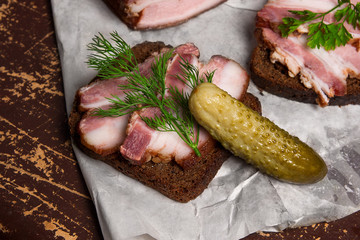 Slices of smoked bacon with rye black bread and several pickles on the white packaging paper.
