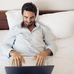 Happy handsome man using laptop. Lying on bed in bedroom.