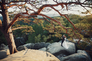 Look from afar at wedding couple hugging on the rocks in evening sun