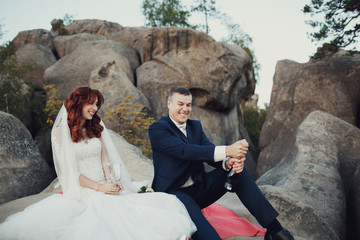 Bride and groom sit on the red blanket on the rocks