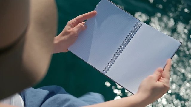 A human being with a hat opens and closes the notebook clean the water. A woman in a wide-brimmed headgear holds a notebook with springs in both of its hands, in which there are no records or drawings
