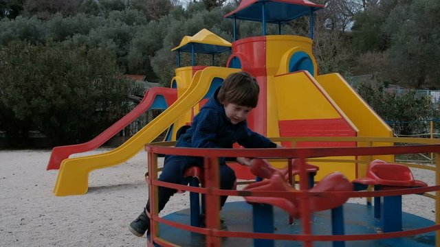 The child quickly spin on the carousel in the playground in the spring. A little boy in a warm blue jacket is played on a round swing with a seat. The kid alone is having fun on a safe attraction. He