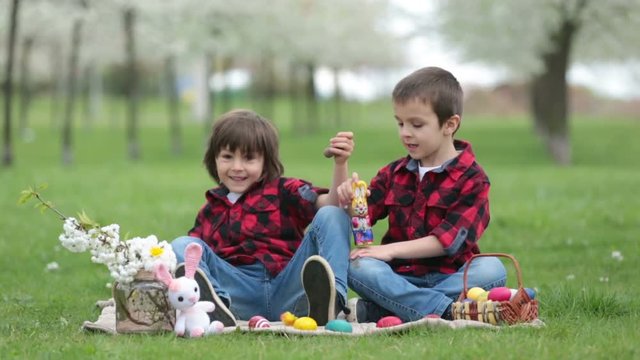 Two children, boy brothers, eating chocolate bunnies and having fun with easter eggs in the park, beautiful spring blooming garden
