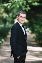 Groom in black suit holds his hands in the pockets posing in park