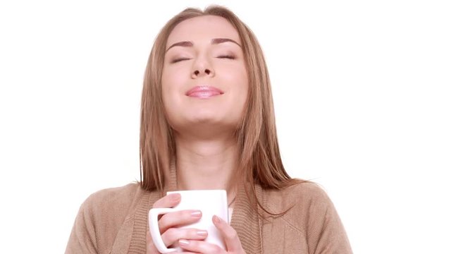 Young beautiful Caucasian female with light brown hair standing on white background drinking hot tea from cup with pleasure