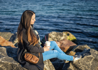 Beautigul girl sitting on the rocky beach with takeaway coffee