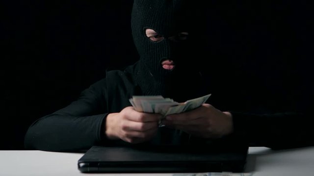 A hacker in a balaclava counts money. Cyber crime.