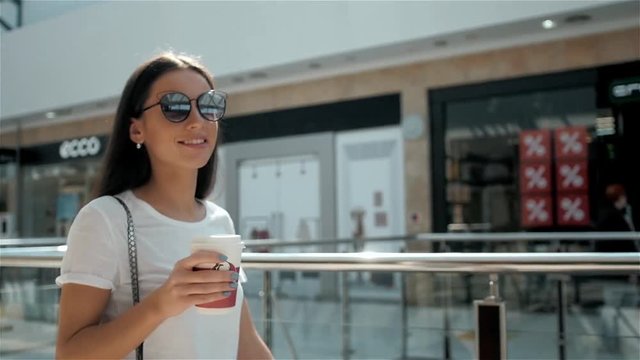Happy young fashionable woman taking a coffee break after shopping, smiling with a coffee-to-go in her hands against shopping mall