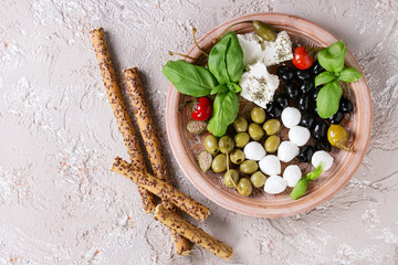 Mediterranean appetizer antipasti plate with green black olives, feta cheese, mozzarella, capers, pepper, basil with grissini bread sticks over beige concrete texture background. Top view with space