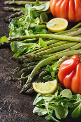 Bundle of young raw uncooked organic green asparagus with green salad leaves, sliced lemon, tomatoes and sea salt over brown texture background. Close up. Healthy eating