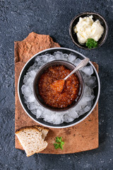 Bowl of red caviar on ice with spoon served with sliced bread, butter and herbs on terracotta board over black stone texture background. Top view with space