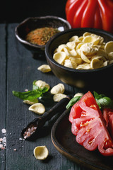 Homemade raw uncooked pasta with whole and sliced organic tomatoes Coeur De Boeuf, salt, seasoning, olive oil and basil over dark wooden background. Italian cuisine