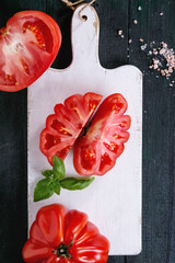 Whole and sliced organic tomatoes Coeur De Boeuf. Beefsteak tomato with pink salt and basil on white wooden chopping board over dark wood background. Top view with space.