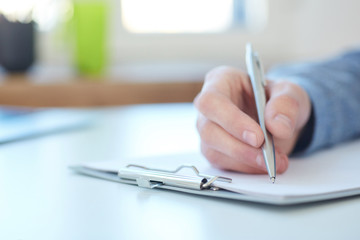 Male hand holding silver pen ready to make note in a clipboard. Businessman or employee at workplace writing business ideas, plans or tasks. Office life or education concept.