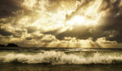 Obraz premium Dramatic cloudscape over the sea with rays of sunlight and a foaming wave in the foreground, toned warm colors