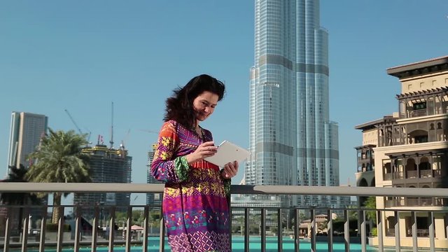 Woman stands near skyscraper and uses tablet computer. Businesswoman with white tablet computer. Adult woman uses tablet computer. Burj Khalifa megatall skyscraper in Dubai, United Arab Emirates