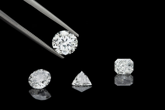 loose brilliant diamonds, one is being held by a tweezers on reflective black background