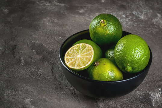 Fresh organic tropical citrus fruits lime in a black bowl on a gray concrete background. Whole and half. Copy space