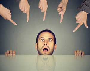 Scared man employee hiding under the table being accused by people who point fingers at him