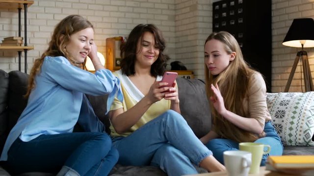 three young and energetic best friend girls look over photos in social network using telephone mobile and discuss emotionally, laughing and impressing surprise sitting on sofa at home during sunny day